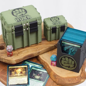 May include: Two olive green storage cases with black latches and The Incredible Hulk fist logos, a black card deck box with a matching logo, a stack of blue-edged trading cards, and two multi-colored polyhedral dice arranged on wooden surfaces.