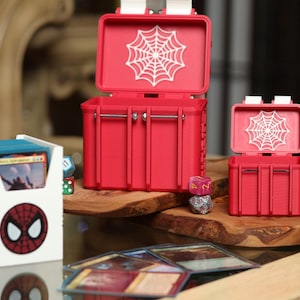 Könnte beinhalten: Zwei rote Plastik-Aufbewahrungsboxen mit einem weißen Spinnennetz-Design auf dem Deckel. Die Boxen sind geöffnet und zeigen das Innere. Die größere Box steht auf einer Holzoberfläche mit einer kleineren Box davor. Auf der Oberfläche befinden sich auch einige Würfel und Spielkarten.