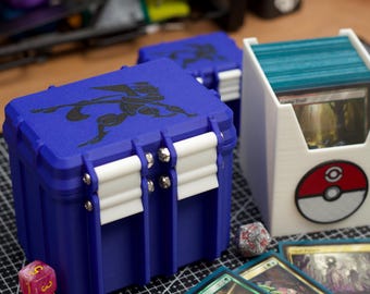 May include: A blue 3D-printed deck box with a black graphic on the lid and white latches, accompanied by a white open-top card holder featuring a red and white circular emblem, alongside several trading cards and multi-sided gaming dice.