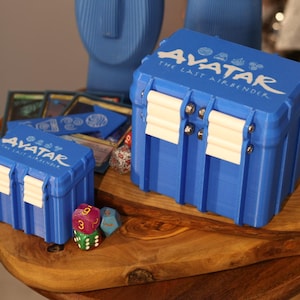 Puede incluir: Dos cajas de almacenamiento azules con el texto "Avatar: The Last Airbender". Las cajas tienen cierres blancos y están sobre una superficie de madera. También se ven dados y cartas.