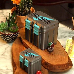 May include: Two grey storage boxes, each with "FINAL FANTASY" in blue and black. The boxes have teal accents and sit on a wooden board. A small dice is next to the boxes. These boxes are likely for storing collectibles.