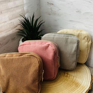 May include: Four corduroy cosmetic bags in shades of brown, pink, gray, and cream, each with a gold zipper. The bags are arranged on a wooden slice, with a small potted plant in the background. The bags are rectangular.
