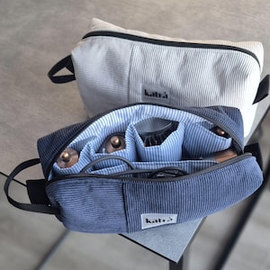 Op de afbeelding: Twee cosmetische tassen van corduroy. De voorste tas is open en onthult compartimenten en schoonheidstools. De tas is marineblauw met een zwarte ritssluiting en handvat. De binnenkant is blauw en wit gestreept. De tweede tas is lichtgrijs.