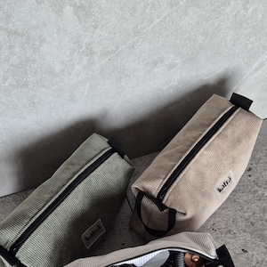 K&ouml;nnte beinhalten: Drei Rei&szlig;verschlusstaschen aus Cord in verschiedenen Farben: Gr&uuml;n, Beige und Wei&szlig;. Die Taschen haben schwarze Rei&szlig;verschl&uuml;sse und schwarze Griffe. Die wei&szlig;e Tasche ist offen und enth&auml;lt einen Haartrockner, eine Rundb&uuml;rste und andere Haarstyling-Werkzeuge. Die Taschen haben den Schriftzug "katla" aufgedruckt.