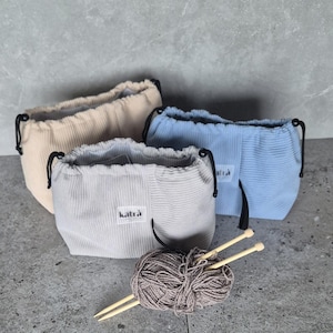 Op de afbeelding: Drie corduroy trekkoordtassen in beige, grijs en blauw, elk met een zwart trekkoord. De grijze tas heeft een wit label met de tekst "katrà". Een bol bruin garen en twee houten breinaalden staan op de voorgrond.