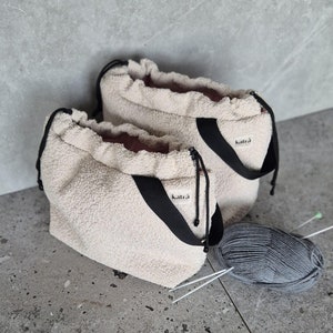 May include: Two cream-colored fuzzy drawstring bags with black straps and the word "katra" printed on the straps. A ball of gray yarn and two knitting needles are on the floor.