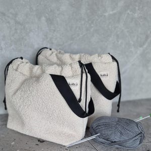 May include: Two white fuzzy drawstring bags with black straps and the word "katra" printed on them. A ball of gray yarn and knitting needles are in the foreground.