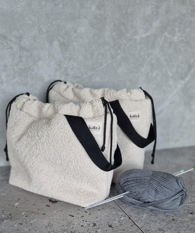 May include: Two white fuzzy drawstring bags with black straps and the word "katra" printed on them. A ball of gray yarn and knitting needles are in the foreground.