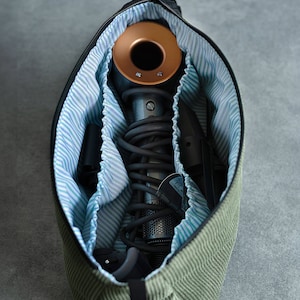 Peut inclure: Un sac de voyage en velours côtelé vert avec une fermeture éclair noire et une sangle noire. Le sac est ouvert et contient un sèche-cheveux, un cordon et un outil de coiffage.