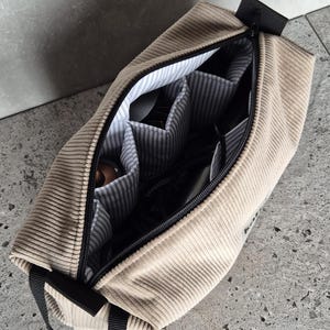 K&ouml;nnte beinhalten: Eine beige Cord-Reisetasche mit schwarzem Rei&szlig;verschluss und Griff. Die Tasche hat ein Rei&szlig;verschlussfach mit mehreren Innentaschen.