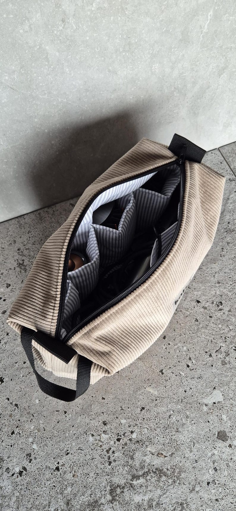 K&ouml;nnte beinhalten: Eine beige Cord-Reisetasche mit schwarzem Rei&szlig;verschluss und Griff. Die Tasche hat ein Rei&szlig;verschlussfach mit mehreren Innentaschen.