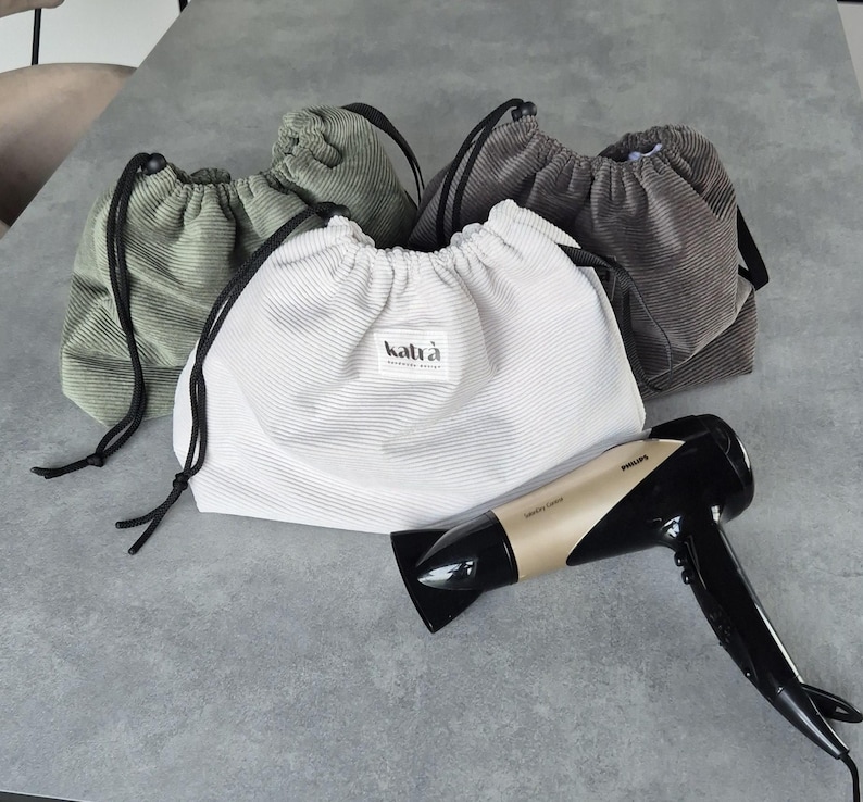 May include: Four drawstring bags made of corduroy fabric in green and brown colors. A white drawstring bag with the text "katr&agrave;" is in the center. A gold hairdryer is lying on the table.