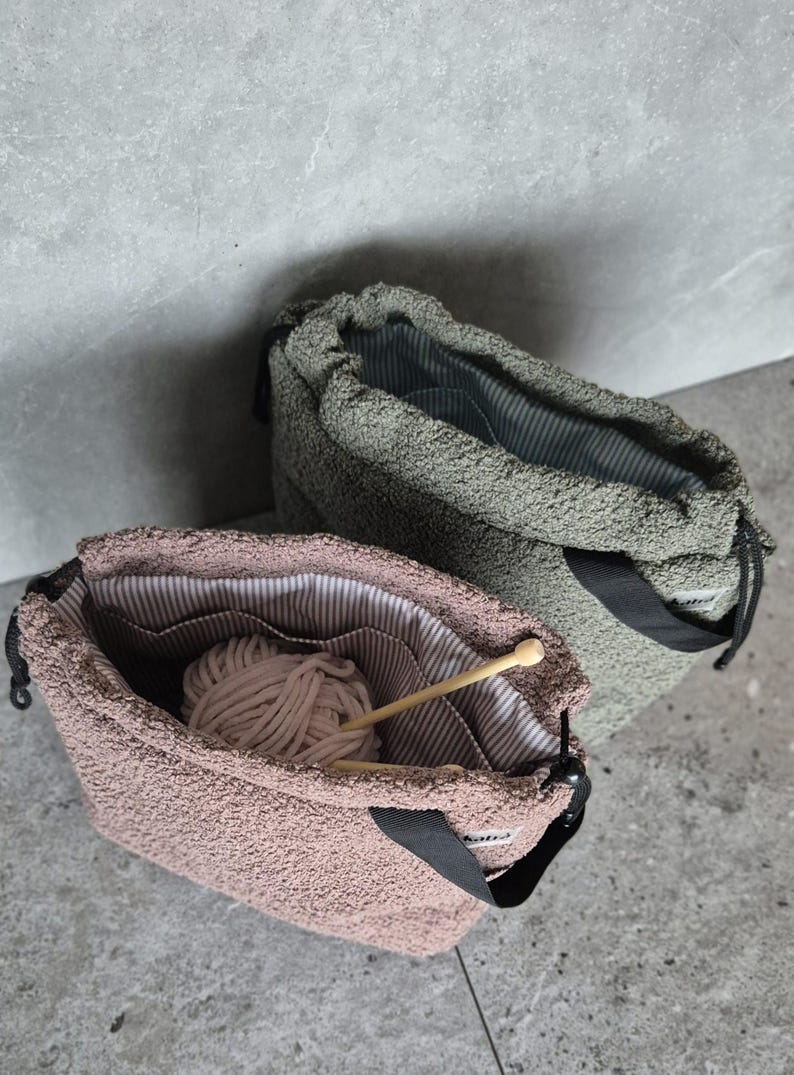 May include: Two drawstring bags made of soft, fuzzy fabric. One bag is green and the other is pink. Both bags have a black strap and a white and black striped lining. The pink bag has a ball of yarn and a wooden knitting needle inside.