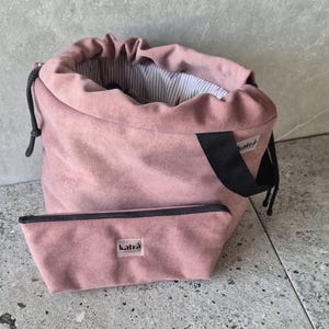 May include: A dusty pink drawstring bucket bag with a black strap and a matching rectangular zippered pouch. Both items feature a small white label with the black text 'katrà'. The bag has a white and grey striped interior lining.