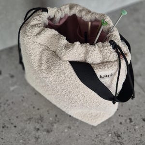 May include: A beige fuzzy drawstring bag with a black strap and the word "katra" printed on the strap. The bag is open and has knitting needles inside.