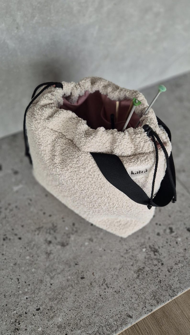 May include: A beige fuzzy drawstring bag with a black strap and the word "katra" printed on the strap. The bag is open and has knitting needles inside.