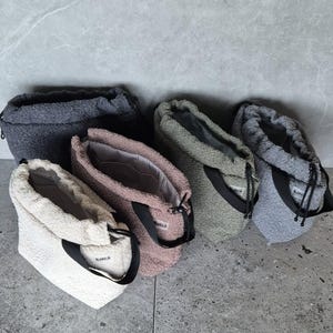 May include: Five fuzzy drawstring bags in different colors, including gray, brown, beige, and green. The bags have black drawstrings and the word "katta" is printed on the bags.