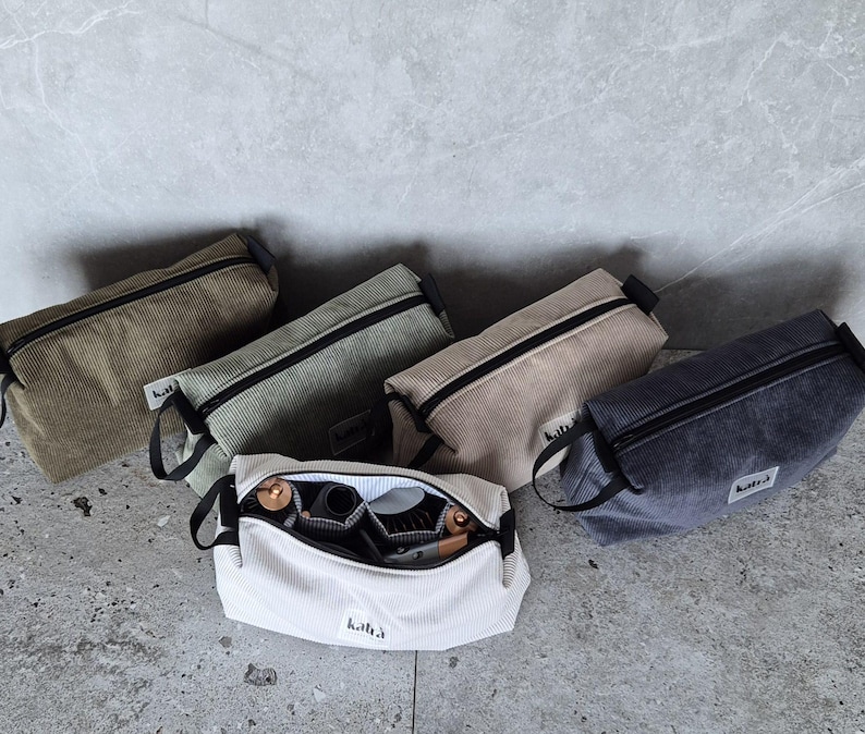 K&ouml;nnte beinhalten: F&uuml;nf verschiedene farbige Cord-Rei&szlig;verschlusstaschen mit schwarzen Rei&szlig;verschl&uuml;ssen. Die Taschen liegen alle auf einer grauen, strukturierten Oberfl&auml;che. Eine Tasche ist wei&szlig; und hat den Schriftzug "kikki.K" aufgedruckt.