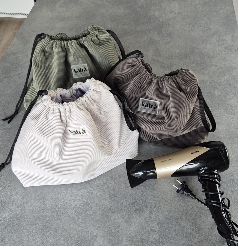 May include: Three drawstring bags made of corduroy fabric in green, brown, and white. The bags have a black drawstring and a label that says "katra". A black and gold hairdryer is also in the image.