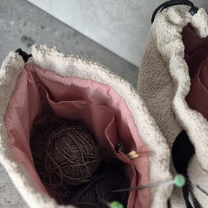 May include: Two beige fuzzy yarn bags with black straps and pink lining. One bag is open and shows a ball of brown yarn and knitting needles.