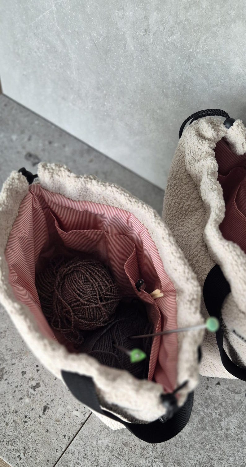 May include: Two beige fuzzy yarn bags with black straps and pink lining. One bag is open and shows a ball of brown yarn and knitting needles.