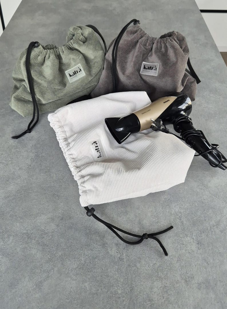 May include: Three drawstring bags made of corduroy fabric in green, gray, and white. The bags have the word "katra" printed on them. A hairdryer is inside the white bag.