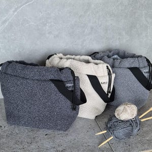 May include: Three fuzzy drawstring bags in gray, white, and light gray with black straps and the word "Knit" printed on the straps. The bags are filled with yarn and knitting needles.