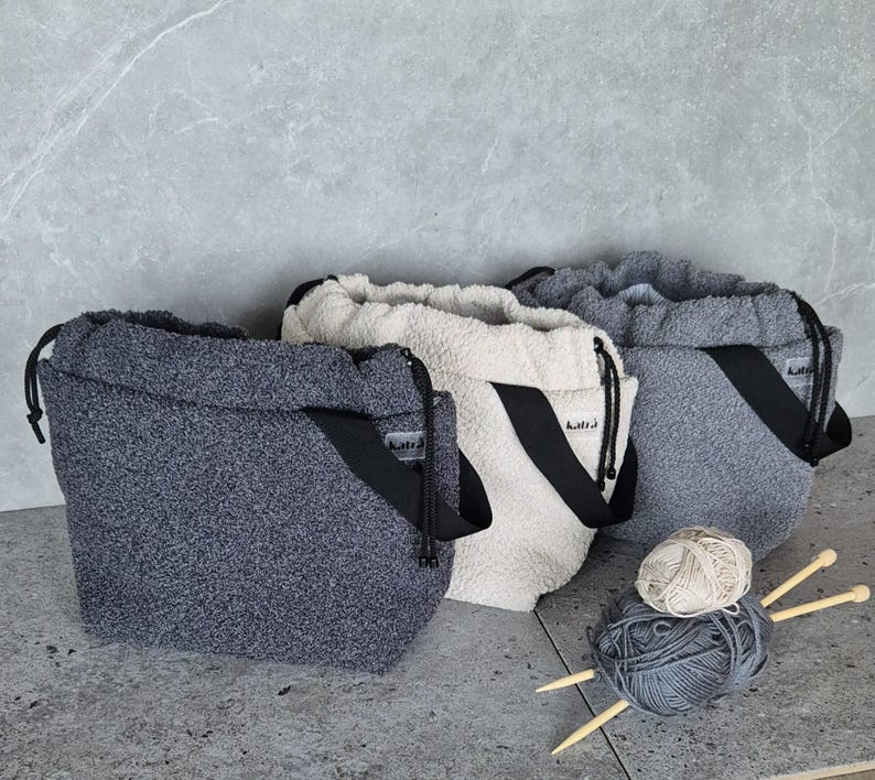 May include: Three fuzzy drawstring bags in gray, white, and light gray with black straps and the word "Knit" printed on the straps. The bags are filled with yarn and knitting needles.