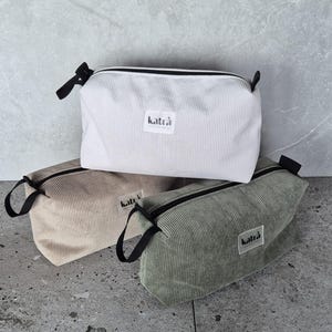 K&ouml;nnte beinhalten: Drei Rei&szlig;verschlusstaschen aus Cordstoff. Die Taschen sind in Wei&szlig;, Beige und Gr&uuml;n. Jede Tasche hat einen schwarzen Rei&szlig;verschluss und ein kleines schwarzes Etikett mit dem aufgedruckten Wort "katra".