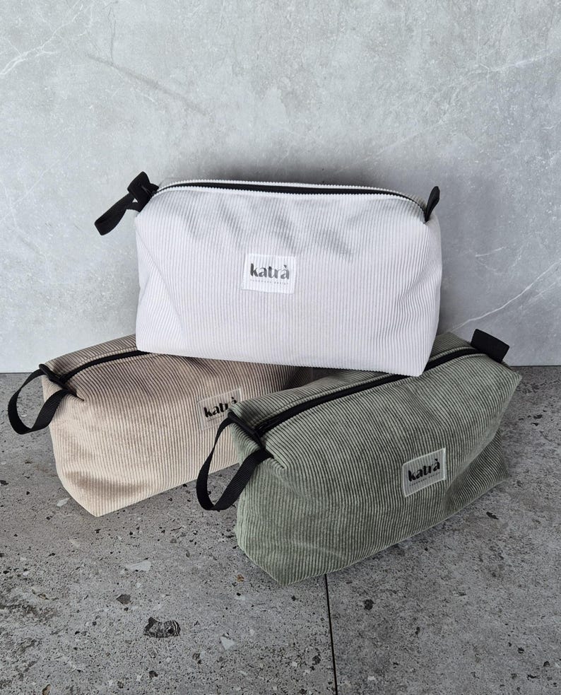 K&ouml;nnte beinhalten: Drei Rei&szlig;verschlusstaschen aus Cordstoff. Die Taschen sind in Wei&szlig;, Beige und Gr&uuml;n. Jede Tasche hat einen schwarzen Rei&szlig;verschluss und ein kleines schwarzes Etikett mit dem aufgedruckten Wort "katra".