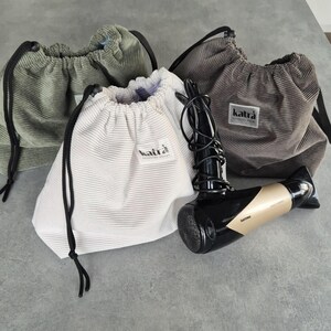 May include: Three drawstring bags made of corduroy fabric in green, white, and gray. Each bag has a black drawstring and a small black label with the word "katra" printed on it. A black and gold hairdryer is lying on the table next to the bags.