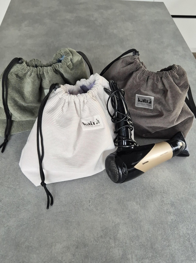 May include: Three drawstring bags made of corduroy fabric in green, white, and gray. Each bag has a black drawstring and a small black label with the word "katra" printed on it. A black and gold hairdryer is lying on the table next to the bags.