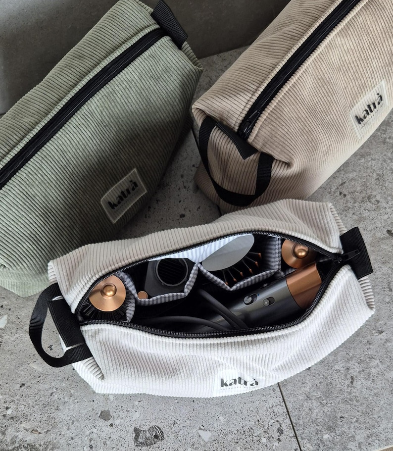 K&ouml;nnte beinhalten: Drei Rei&szlig;verschlusstaschen aus weichem, strukturiertem Stoff. Die Taschen sind in Gr&uuml;n, Beige und Wei&szlig;. Die wei&szlig;e Tasche ist offen und enth&auml;lt einen Haartrockner und andere Styling-Werkzeuge. Die Taschen tragen den Schriftzug "katra".
