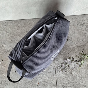 K&ouml;nnte beinhalten: Eine graue Reisetasche aus Cord mit Rei&szlig;verschluss, schwarzem Griff und mehreren Innentaschen. Die Tasche ist ge&ouml;ffnet und das Innere ist mit einem wei&szlig;-grau gestreiften Stoff ausgekleidet.