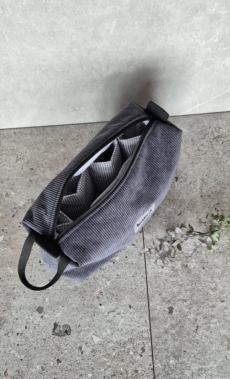 K&ouml;nnte beinhalten: Eine graue Reisetasche aus Cord mit Rei&szlig;verschluss, schwarzem Griff und mehreren Innentaschen. Die Tasche ist ge&ouml;ffnet und das Innere ist mit einem wei&szlig;-grau gestreiften Stoff ausgekleidet.