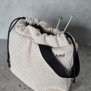 May include: A white fuzzy drawstring bag with a black strap and the word "katra" printed on the front. Two green knitting needles are sticking out of the top of the bag.