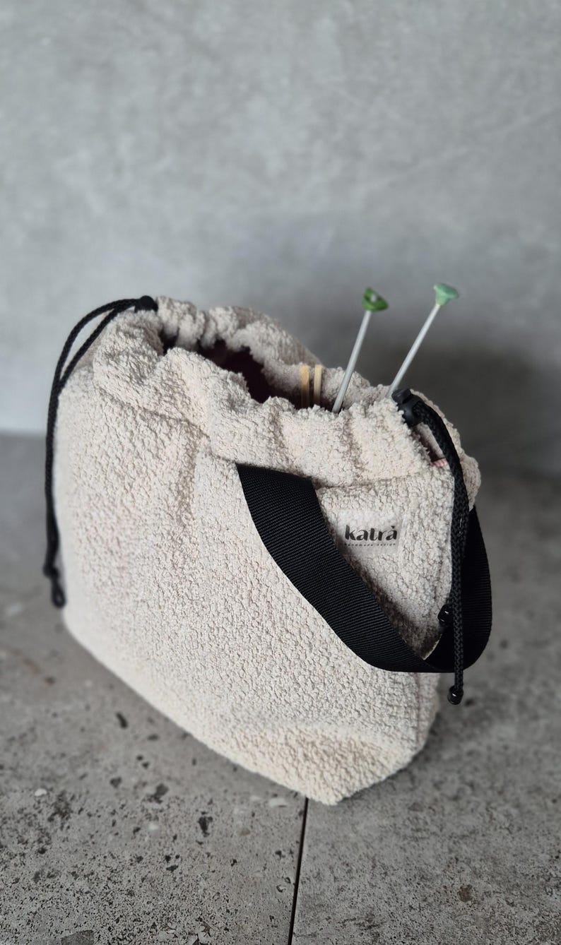 May include: A white fuzzy drawstring bag with a black strap and the word "katra" printed on the front. Two green knitting needles are sticking out of the top of the bag.