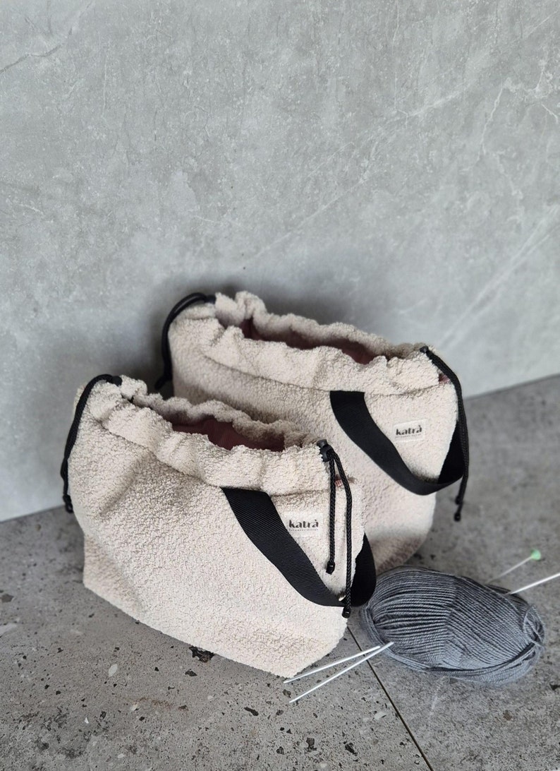 May include: Two cream-colored fuzzy drawstring bags with black straps and the word "katra" printed on the straps. A ball of gray yarn and two knitting needles are on the floor.
