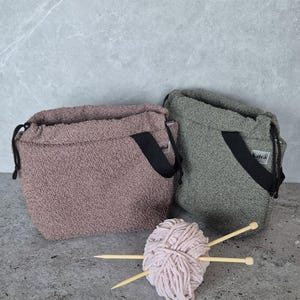 May include: Two fuzzy drawstring bags with black straps, one in a light brown color and the other in a dark green color. A ball of pink yarn and two wooden knitting needles are in front of the bags.
