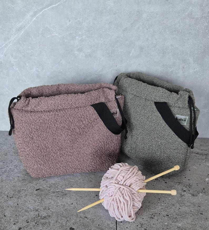 May include: Two fuzzy drawstring bags with black straps, one in a light brown color and the other in a dark green color. A ball of pink yarn and two wooden knitting needles are in front of the bags.