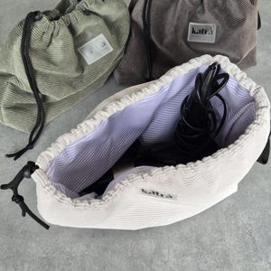 May include: Three drawstring bags made of corduroy fabric. The bags are in green, gray, and white. The white bag is open and has a black hair dryer inside. All bags have a black drawstring and a label that says "katra".