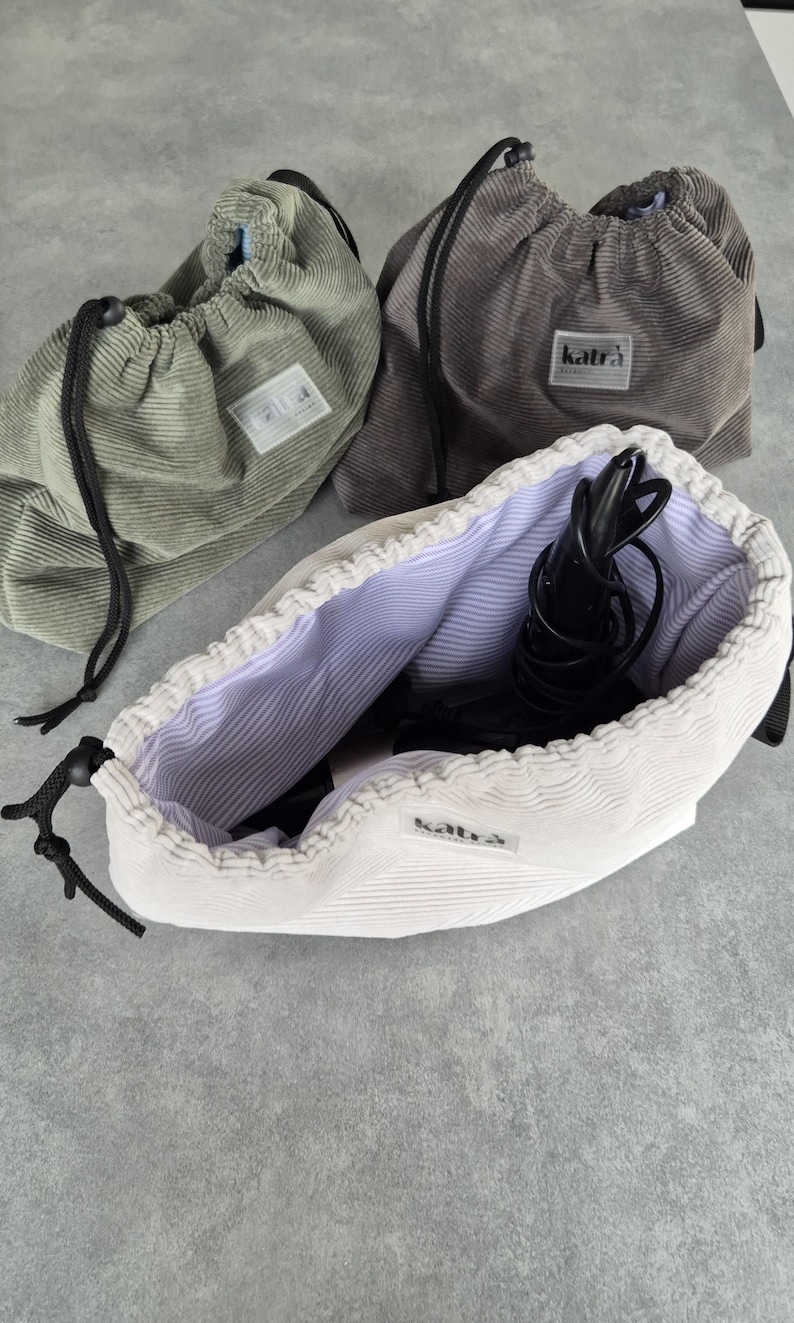 May include: Three drawstring bags made of corduroy fabric. The bags are in green, gray, and white. The white bag is open and has a black hair dryer inside. All bags have a black drawstring and a label that says "katra".