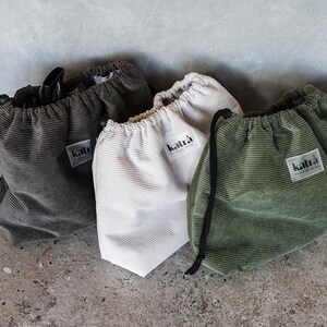 May include: Three drawstring bags made of corduroy fabric in green, white, and gray. Each bag has a label with the word "katra" printed on it.