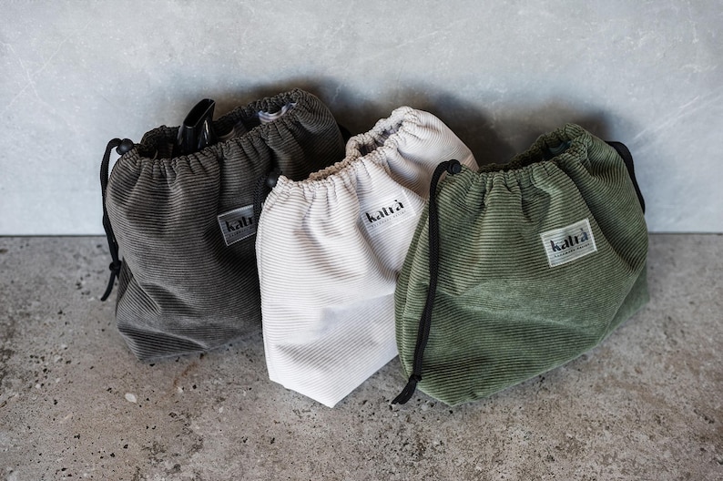 May include: Three drawstring bags made of corduroy fabric in green, white, and gray. Each bag has a label with the word "katra" printed on it.