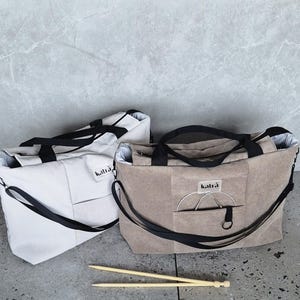 May include: Two beige corduroy tote bags with black straps and the word "katra" printed on the front. The bags are sitting on a light grey surface with two wooden knitting needles.