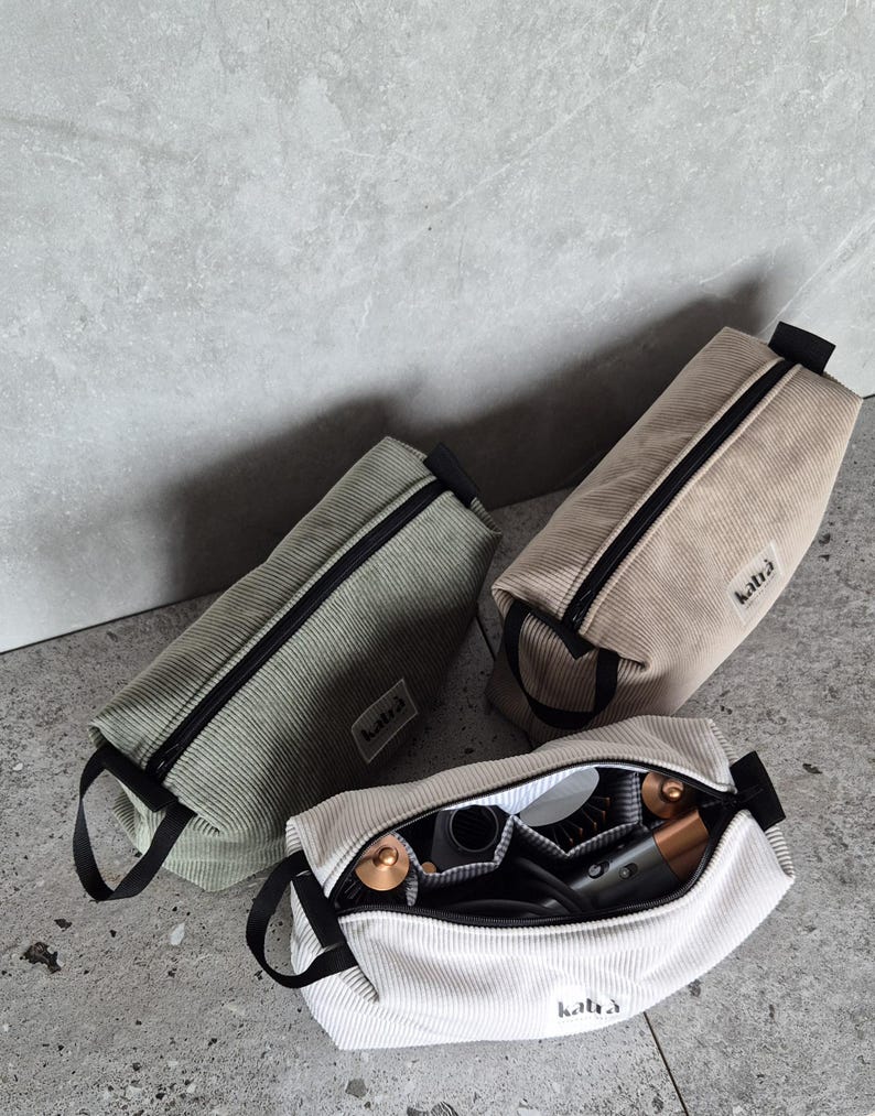K&ouml;nnte beinhalten: Drei Rei&szlig;verschlusstaschen aus Cord in verschiedenen Farben: Gr&uuml;n, Beige und Wei&szlig;. Die Taschen haben schwarze Rei&szlig;verschl&uuml;sse und schwarze Griffe. Die wei&szlig;e Tasche ist offen und enth&auml;lt einen Haartrockner, eine Rundb&uuml;rste und andere Haarstyling-Werkzeuge. Die Taschen haben den Schriftzug "katla" aufgedruckt.