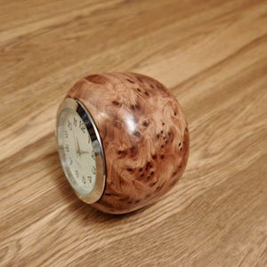May include: A round wooden clock with a silver face and black hands. The clock is made of burl wood with a unique grain pattern.