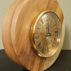 May include: A wooden clock with a gold rim and Roman numeral face. The clock is sitting on a dark surface with two coins in front of it.