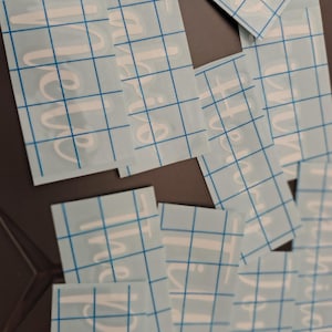 Könnte beinhalten: Eine Ansammlung weißer Vinyl-Aufkleber mit dem Wort "Thankful" in Schreibschrift. Jeder Aufkleber befindet sich auf einer hellblauen Rückseite mit einem Rastermuster. Die Aufkleber sind auf einer dunklen Oberfläche verstreut.