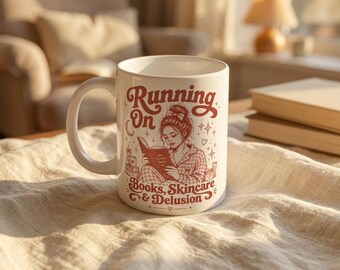 May include: White ceramic mug with a wraparound design featuring a woman reading a book, with the text "Running On Books, Skincare & Delusion" in red. The mug has a curved handle and is sitting on a textured surface.